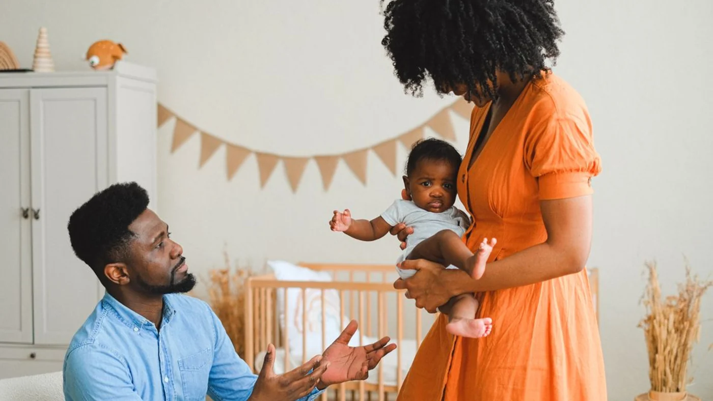 Two caregivers talk in a nursery while one holds the baby; the crib in the background is empty.