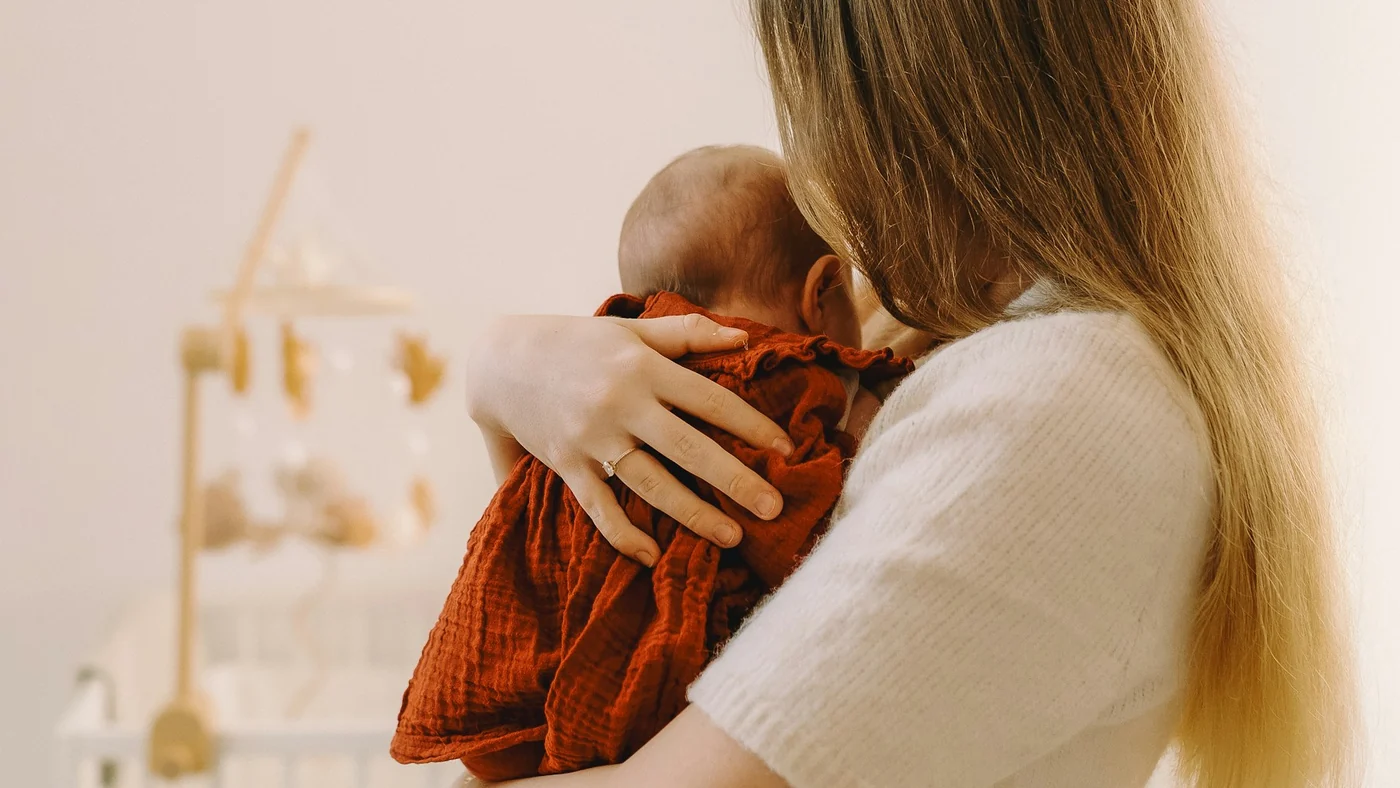 A caregiver holds a baby close in a bright room, with a real crib blurred in the background.